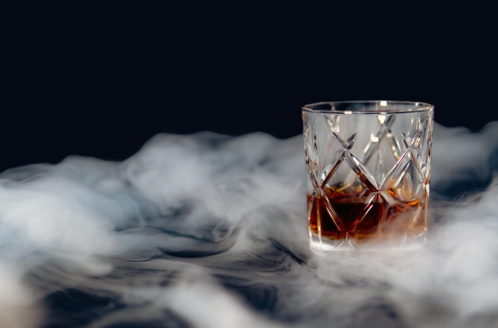 A glass of whiskey on a table covered with smoke against a black background